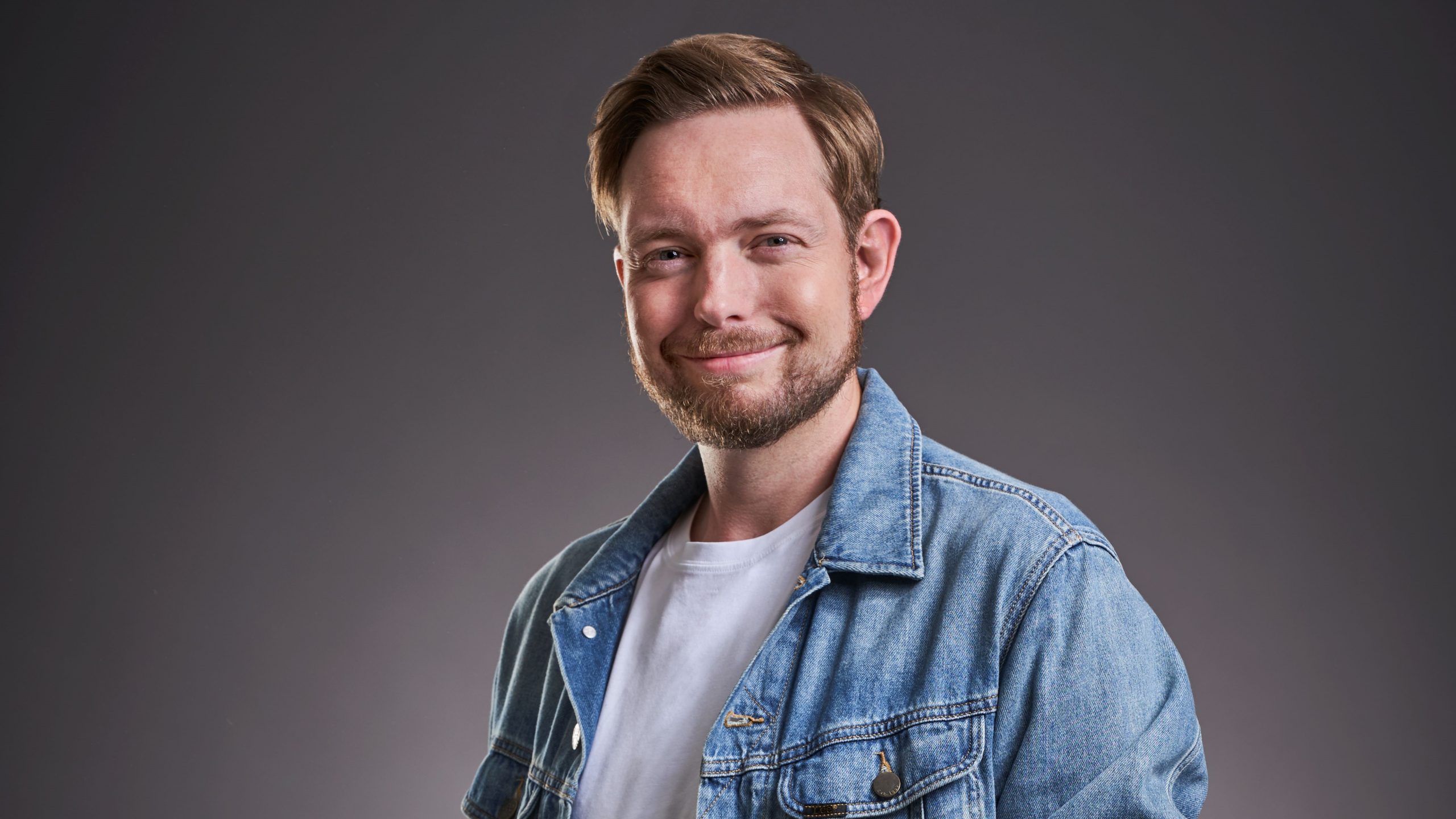 Ein Mann mit kurzen hellbraunen Haaren und Bart lächelt in die Kamera. Er trägt ein weißes T-Shirt unter einer hellblauen Jeansjacke. Der Hintergrund ist dunkelgrau.